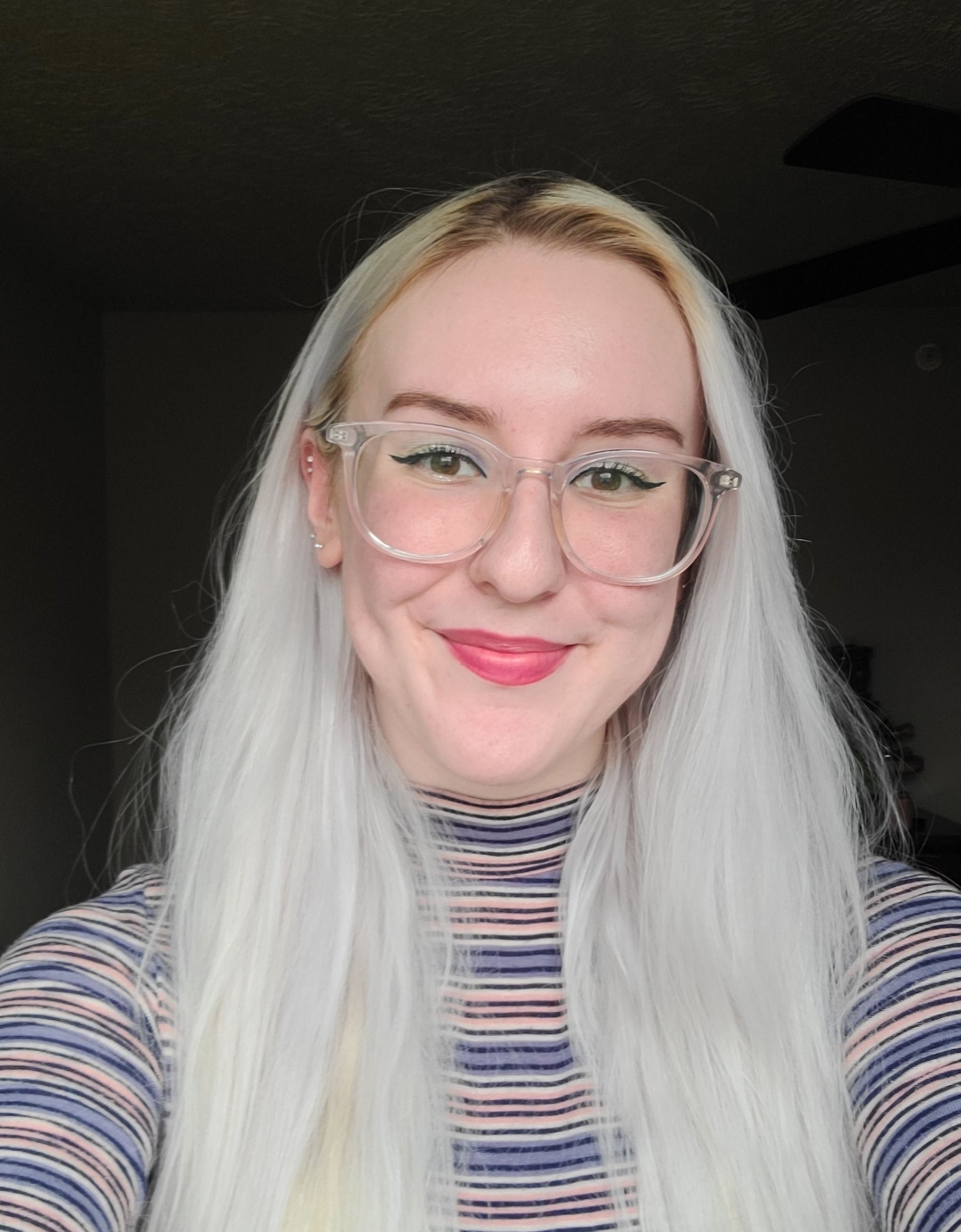 platinum blonde white woman with clear frame glasses smiling wearing a colorful stripped turtle neck shirt