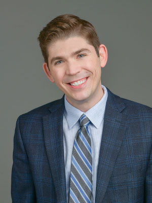 White man, blue eyes, brown hair, wearing a dark blue suite jacket, light blue dress shirt, blue and grey tie